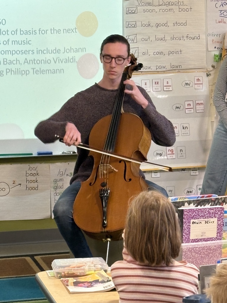 A high school student plays a cello in a classroom, with a projector screen behind him showing music composers. A student sits in front.