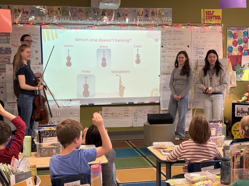 Classroom with a projector screen. Four adults stand, and a violin is held. Students sit at desks, engaged.