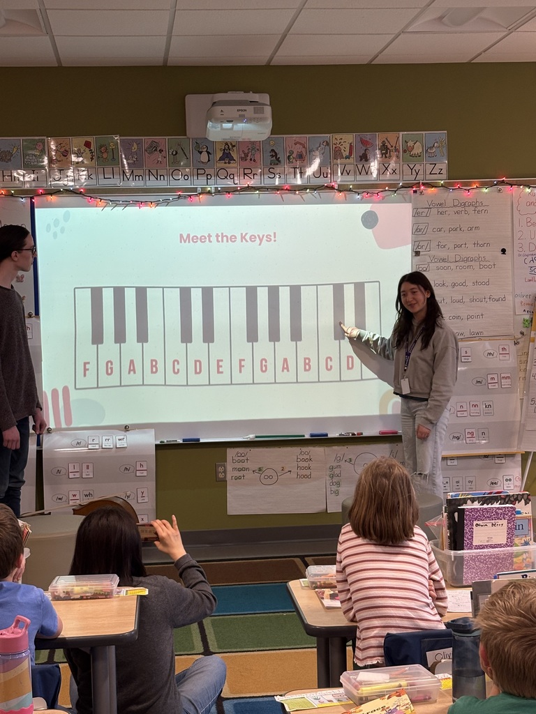 A high school student shows a piano keyboard on a screen in a classroom. Students sit at desks, watching the presentation.