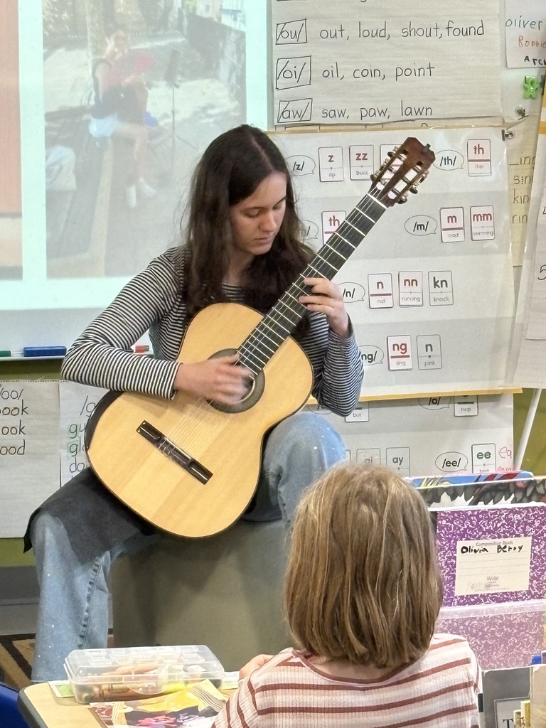 A high school student sitting on stool with guitar, performing in front of a student in a classroom.