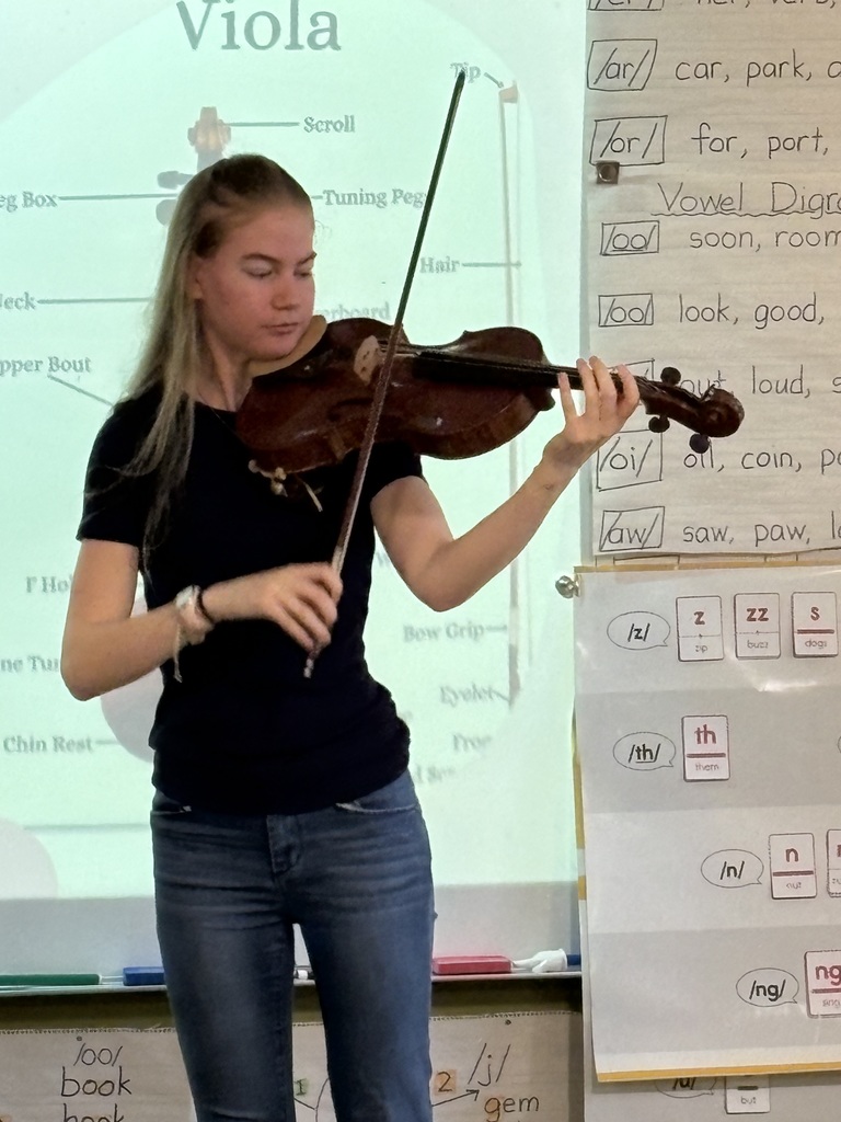 A high school student in a black t-shirt and blue jeans is playing a violin in front of a whiteboard.