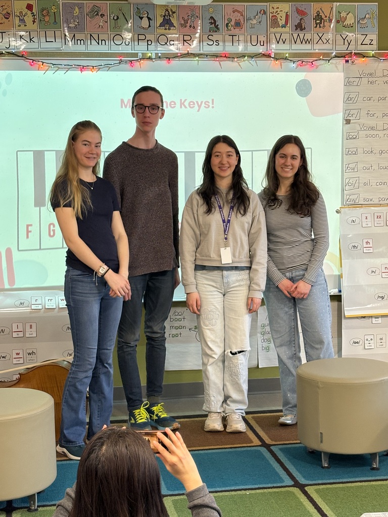 Four high school musicians stand together in front of a screen with a piano keyboard, a teacher takes a picture.