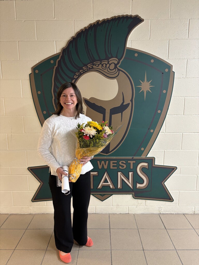 A woman smiles in front of a white wall with a green sign reading "West Titans". She holds a bouquet of flowers.