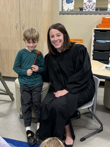 Woman in a black robe and a boy holding a mallet sit together in a classroom.