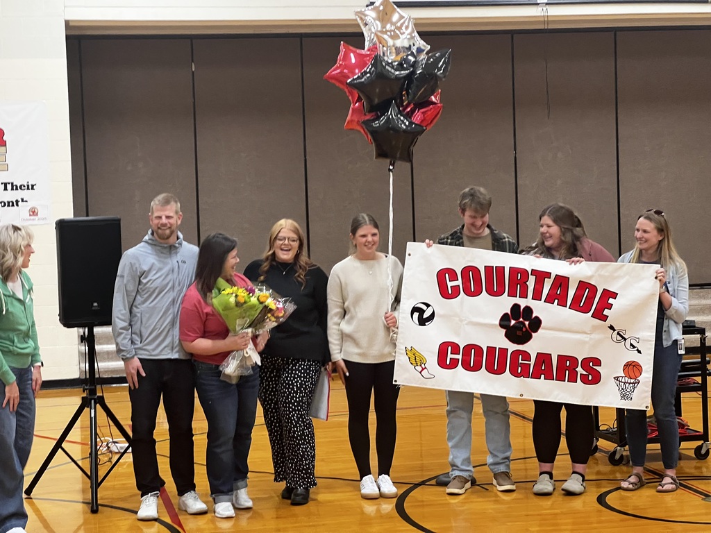 A group of people stands on a basketball court holding a banner that reads "Courtade Cougars". They are smiling.
