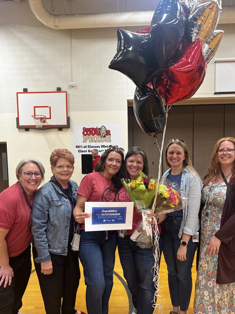 A group of women stand in a gymnasium with balloons. A central woman holds a bouquet and certificate. A basketball hoop is in the background.