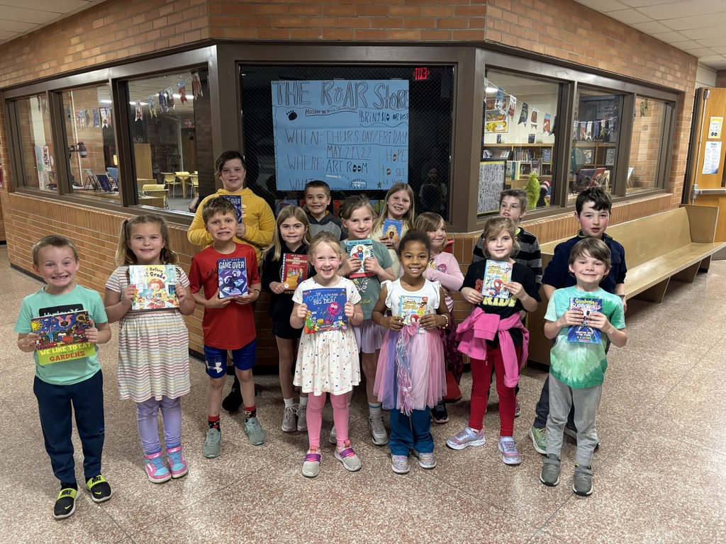 Group of children in a hallway with a display board and bench in the background, holding books.