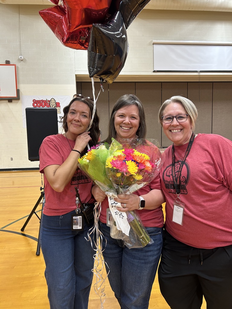 Three women in pink shirts hold a bouquet of flowers and balloons, standing in a gymnasium.