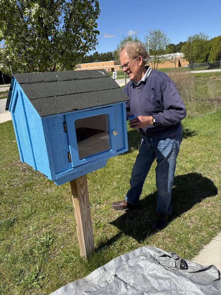 Person standing on grass is painting a blue free library. Trees and building in background. Person wearing glasses and a watch.