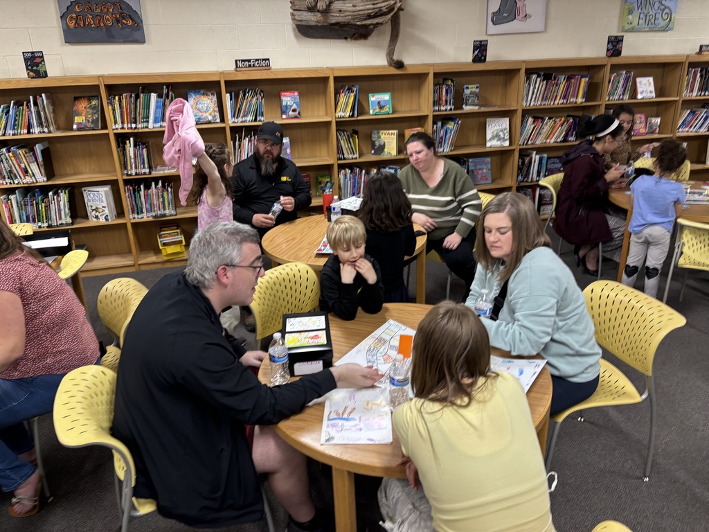 People sit around tables in a library. A man points to a map. Shelves filled with books are in the background.