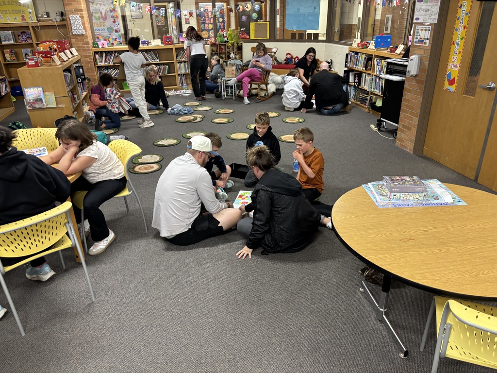 People sitting on floor in a room with chairs, tables, books, and a door in the background.
