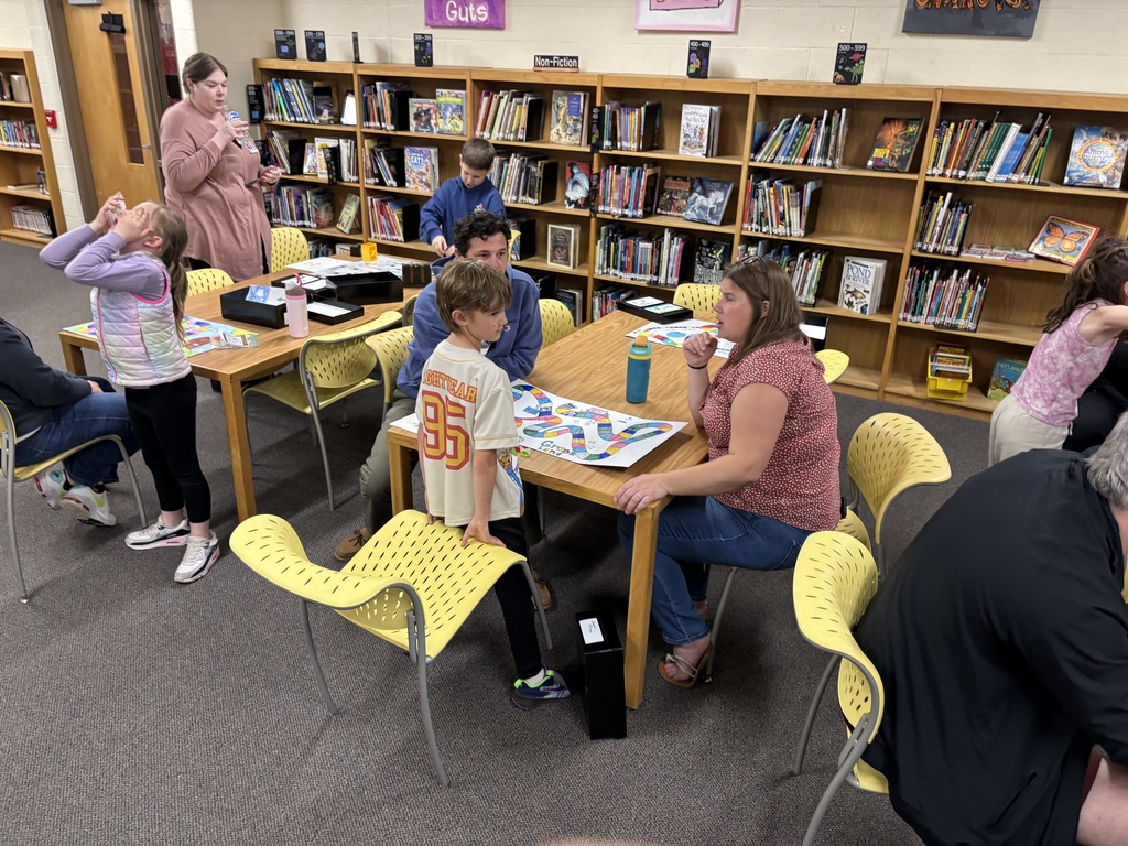 Library room with shelves of books. A group of adults and children playing a board game.