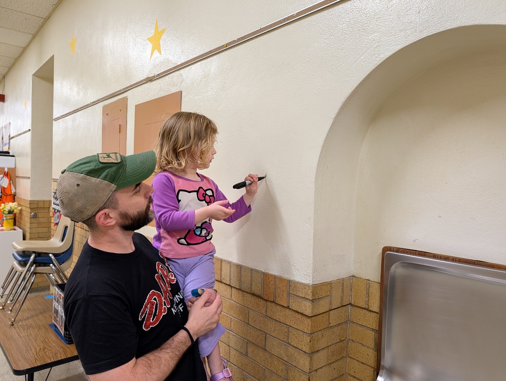 A man holds a young girl up against a wall. The girl draws on the wall with a marker.