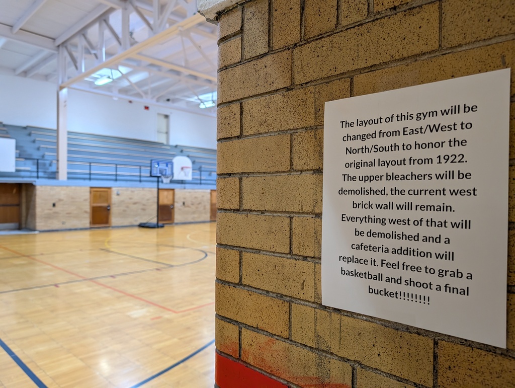 Indoor basketball court with a brick wall. A sign on the wall explains the layout changes to honor a historic date.