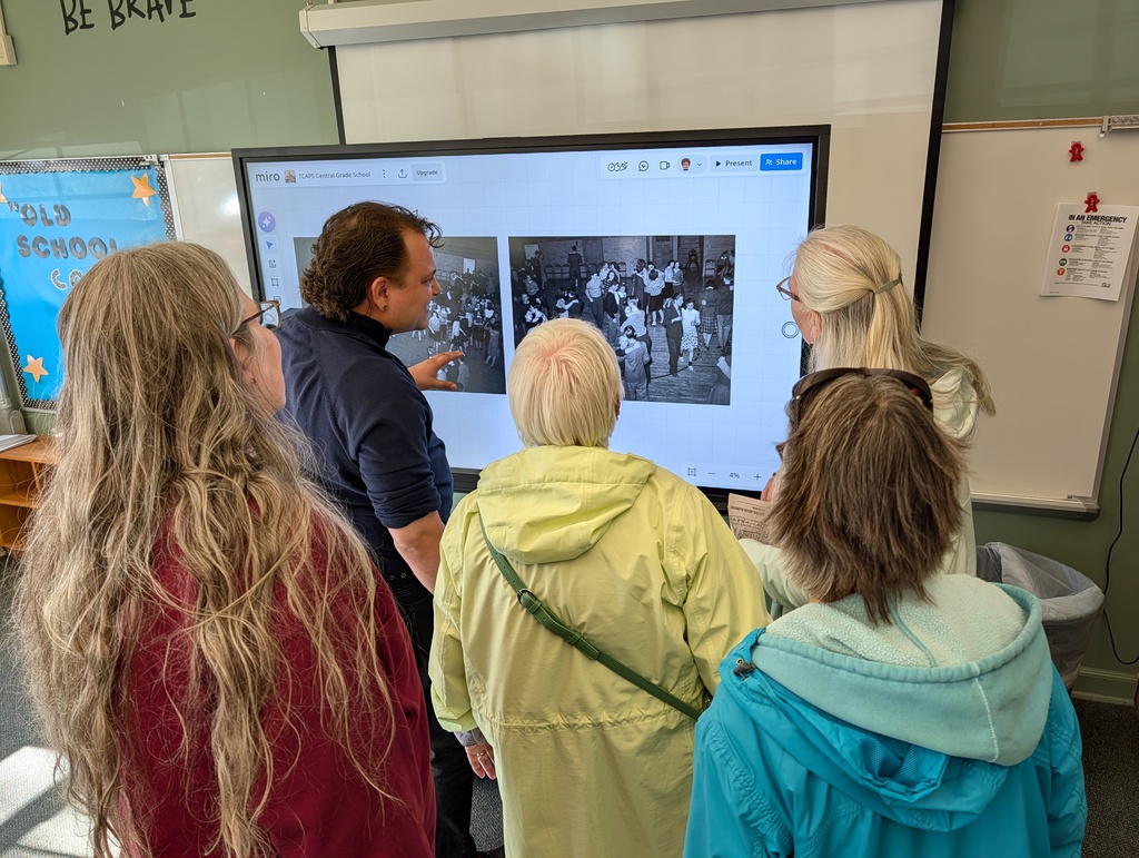 Five adults, one man and four women, watch a black-and-white image on a classroom monitor.