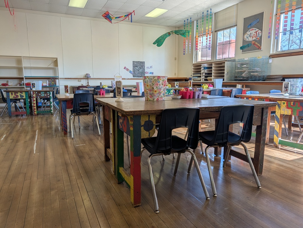 An empty classroom with wooden floors, white walls, and tables with chairs. Colorful decorations are on the walls.