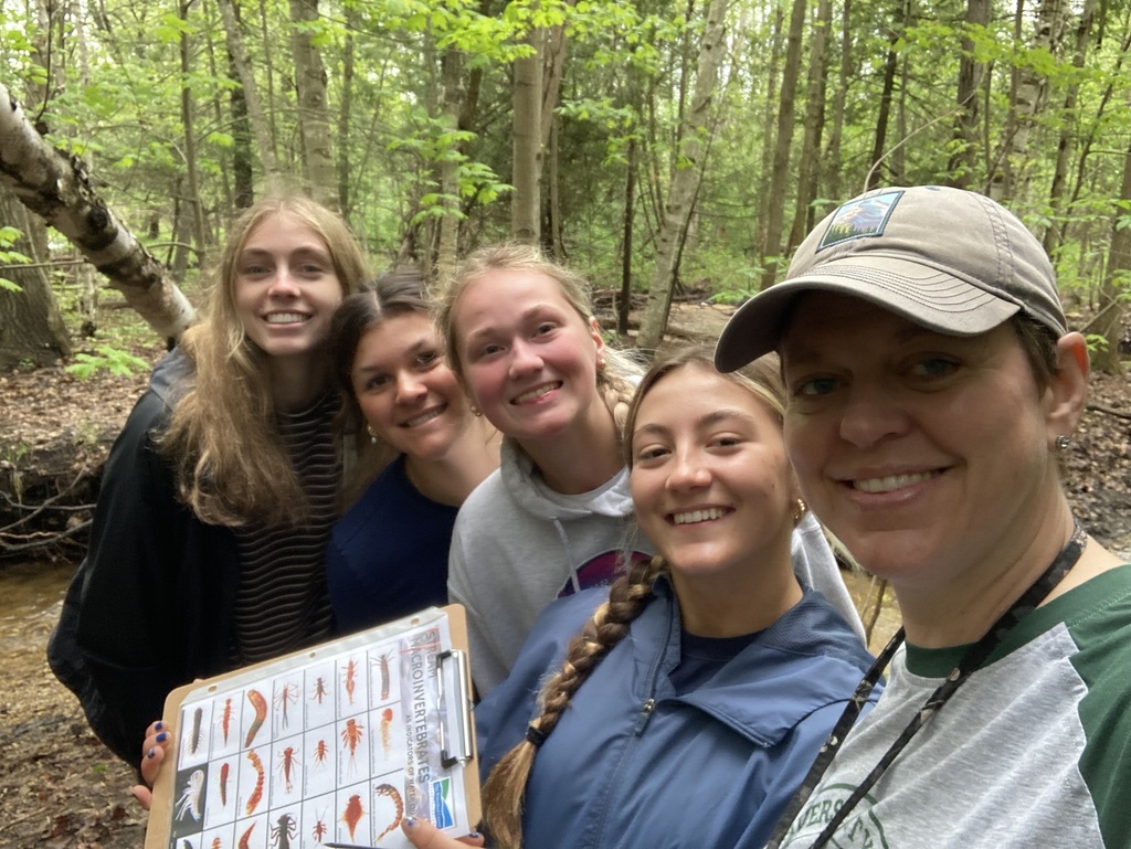 Five women in a forest smile at the camera. One woman holds a clipboard with a grid of small holes.