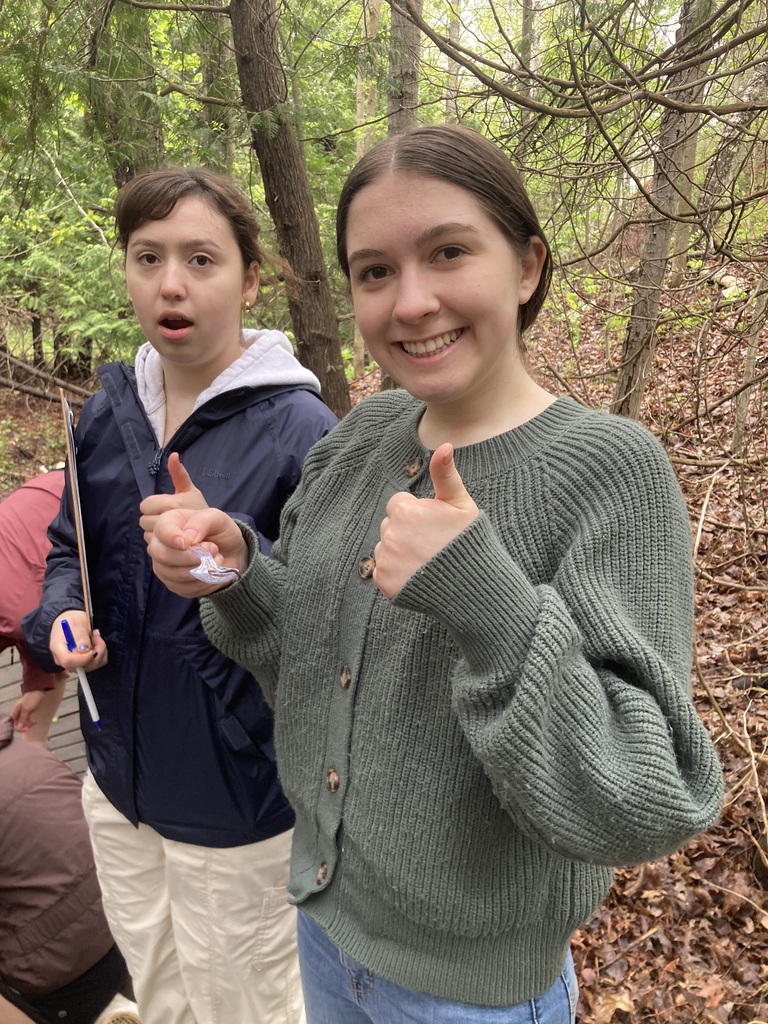 Two people stand in a forest, smiling and giving thumbs up. One holds a pen. Behind them, another person sits on the ground.