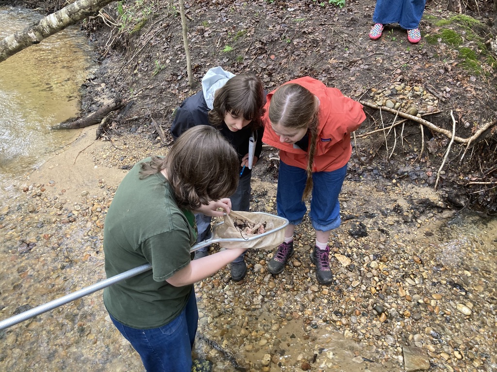 Three people are outdoors, examining a shallow body of water. They are wearing jackets and boots.