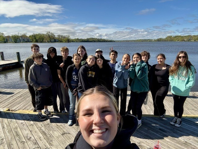 A group of people stands on a dock next to a lake with trees and houses in the background.