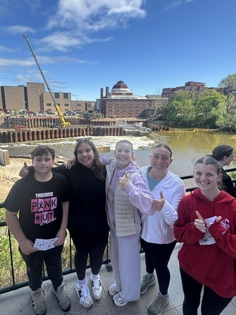 Five young people standing on a railing, smiling, and giving thumbs up. A river and city buildings are behind them.