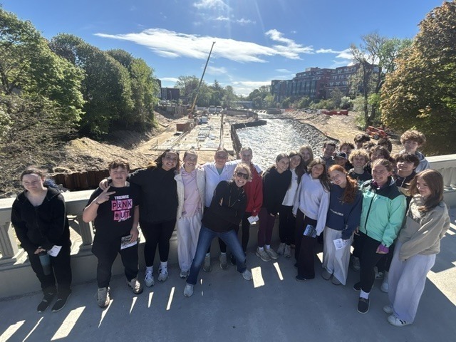 A group of people standing on a bridge over a body of water, with a construction site and greenery in the background.