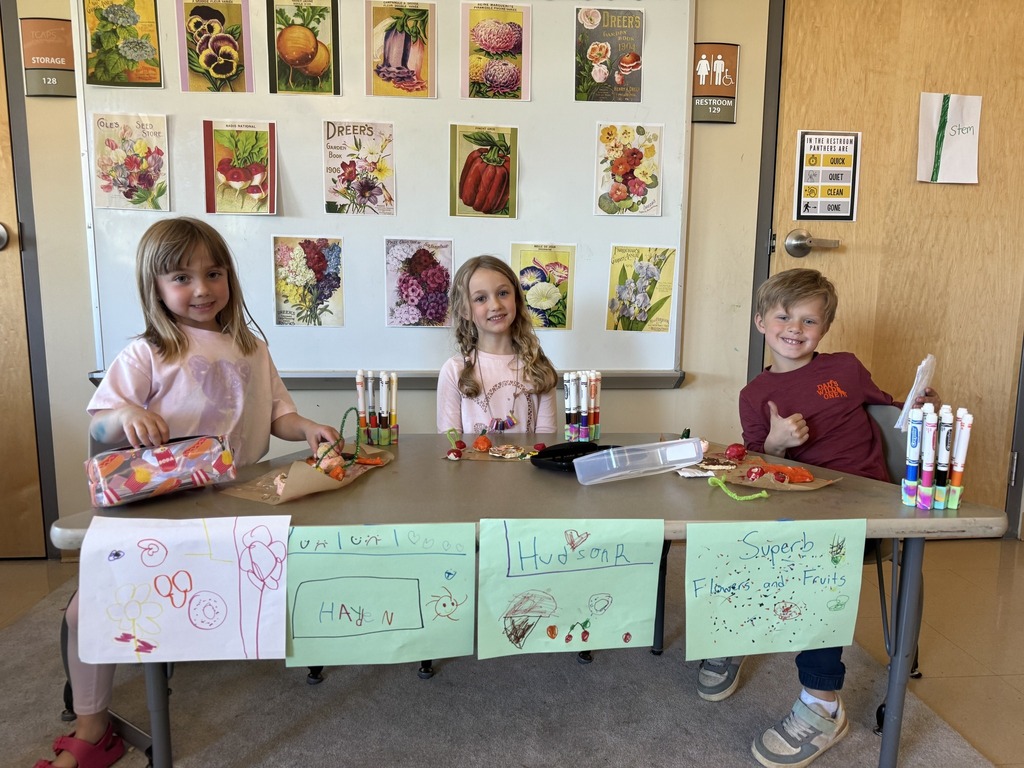 Three children sitting at a table with artwork and various items, against a wall with framed paintings.