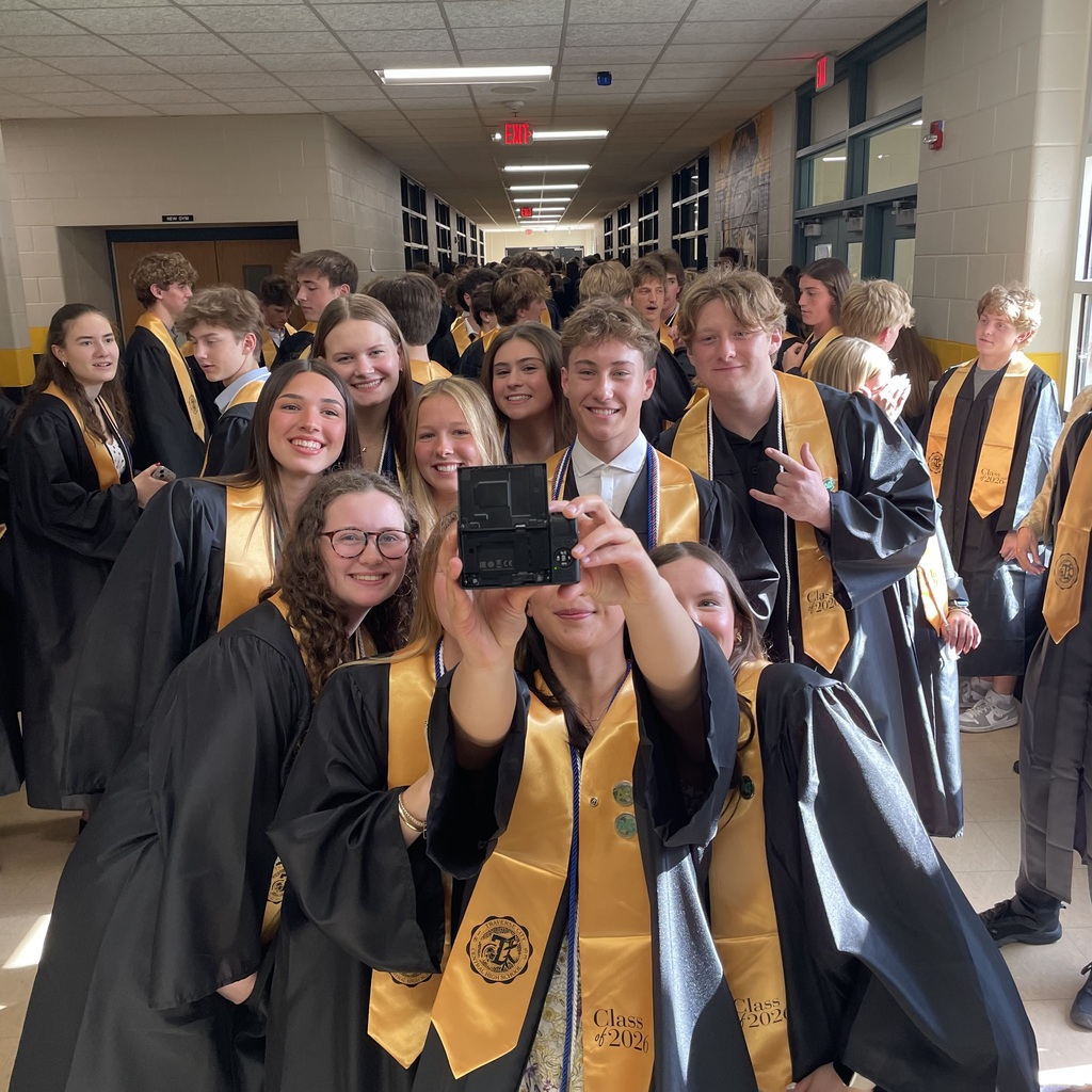 Students in graduation robes gather in a hallway, smiling and posing for a photo with a cellphone.