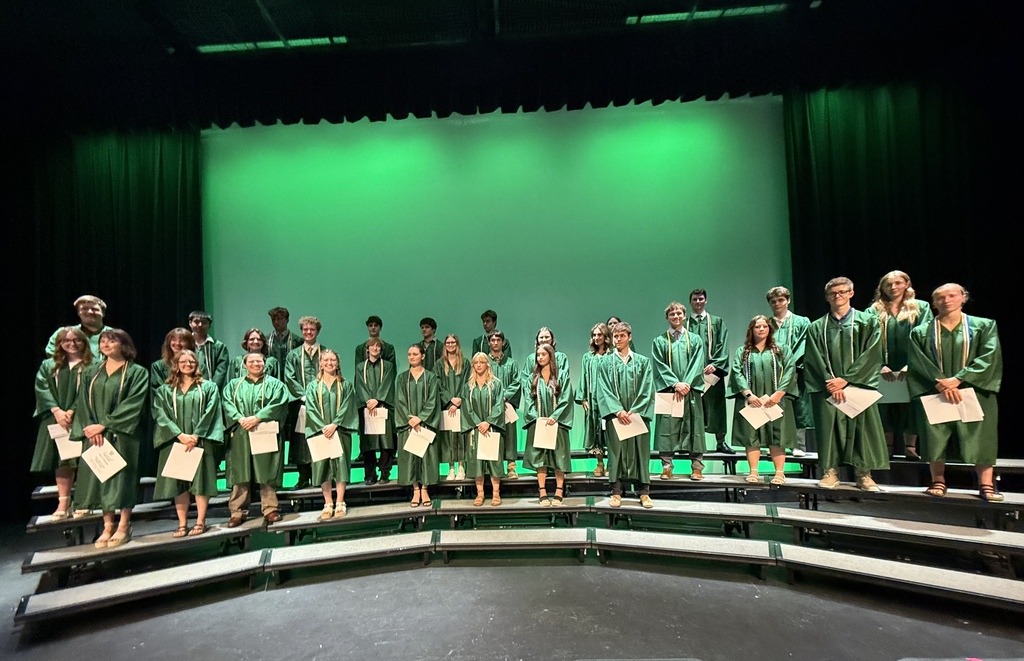 A group of people in green graduation robes stand on a stage, holding papers, with a green screen as the background.