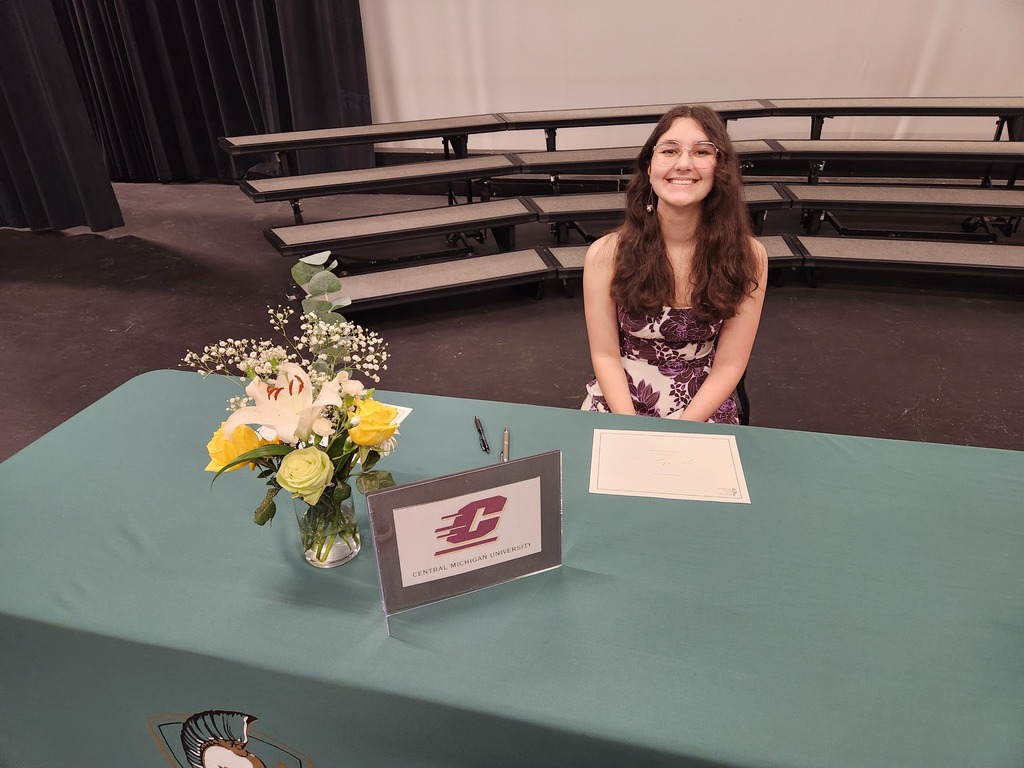 A woman sits at a green table with a flower arrangement, a pen, and a sign. Empty bleachers are in the background.