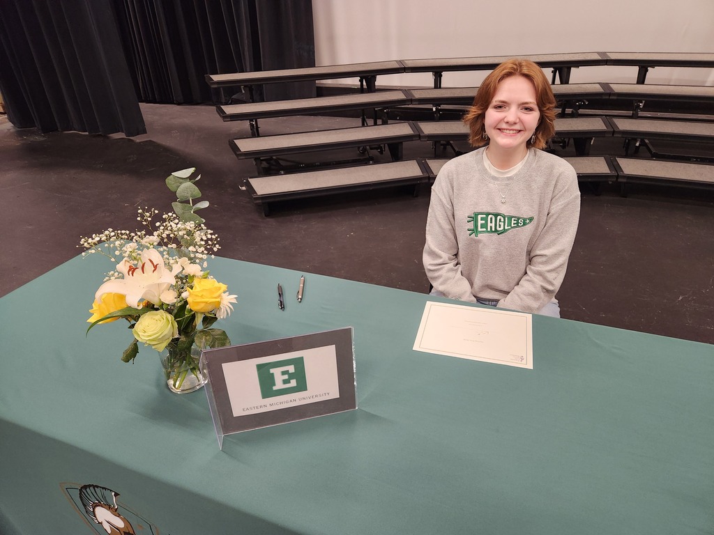 Person standing behind a desk with a sign reading "E" and flowers in a vase.