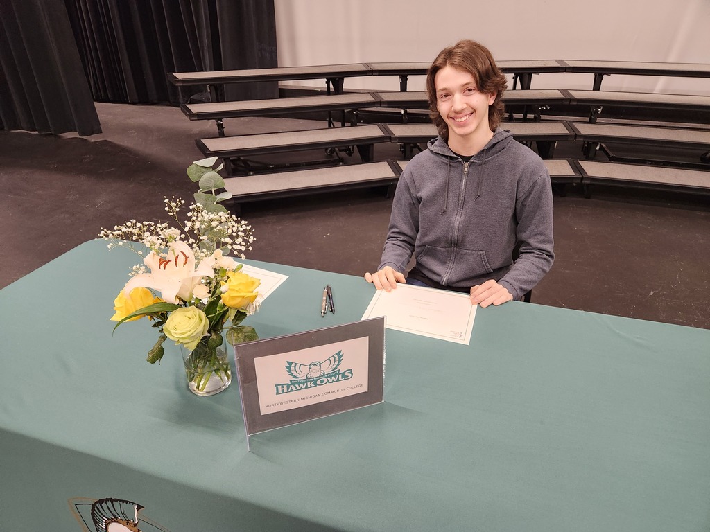 Person seated at a table with papers, pen, and flowers. Behind them is a bleacher seating area.