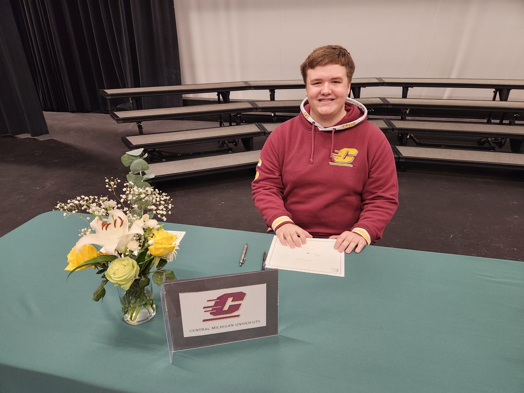 Person at a table with a flower arrangement, holding papers and a pen, in front of an empty bleacher.