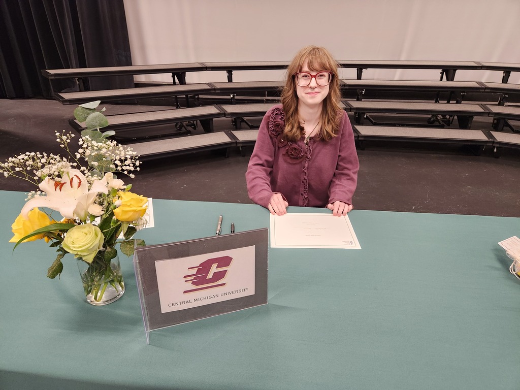 A woman with glasses sits at a desk in a school auditorium, with flowers, a sign, and a paper.