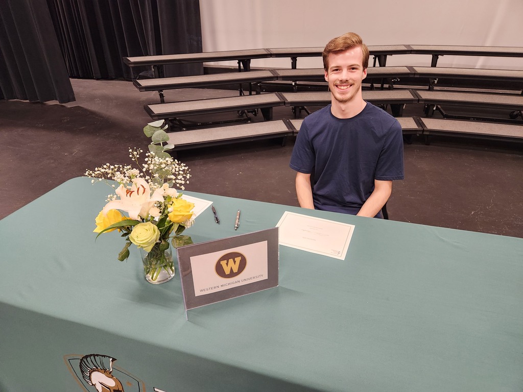 A person in a blue shirt sits at a table in an auditorium with a flower arrangement and a sign.