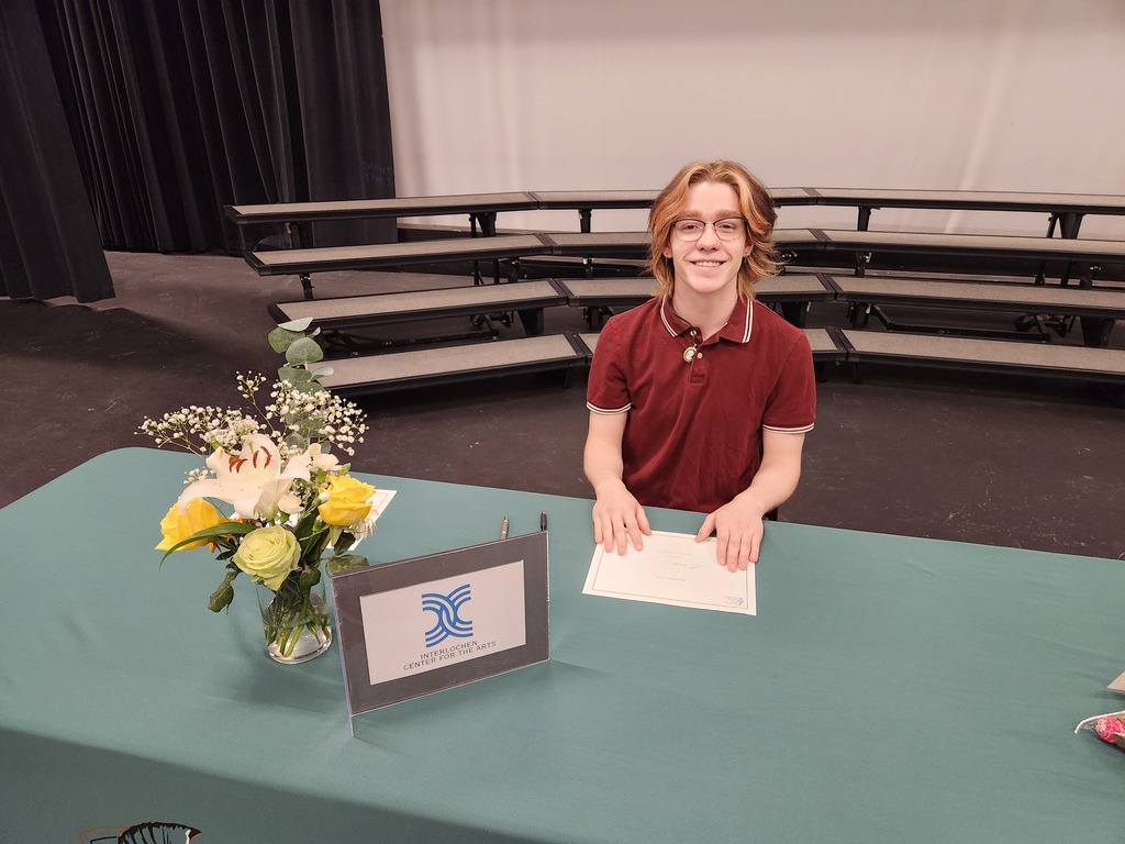 Person in red shirt seated at a table with a flower arrangement and a sign in front.