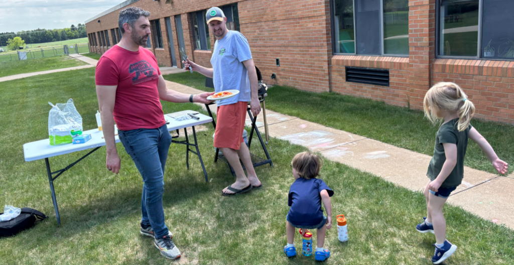 Three adults and two children stand on a lawn. One adult holds a frisbee. A table holds bottles and bags.