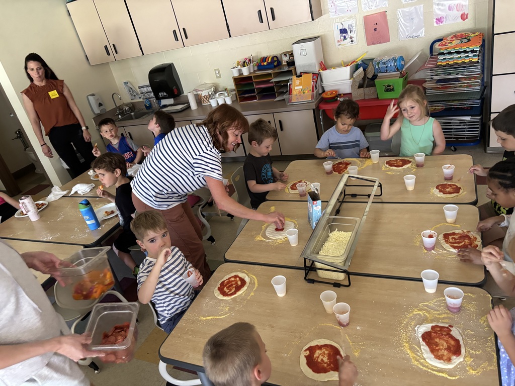 A group of children seated around tables in a classroom, preparing pizzas. An adult helps them.