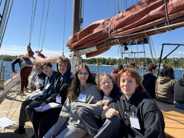 A group of people, some wearing glasses and IDs, are sitting on a deck of a boat in water.