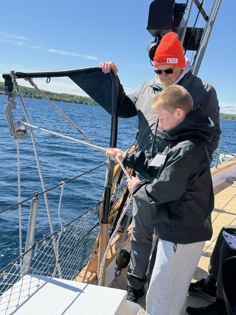 A person wearing a red beanie stands with a child on a boat. Both are holding a rope.