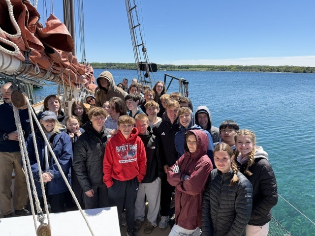 A group of individuals in warm clothing stands on the deck of a boat. The water is calm.