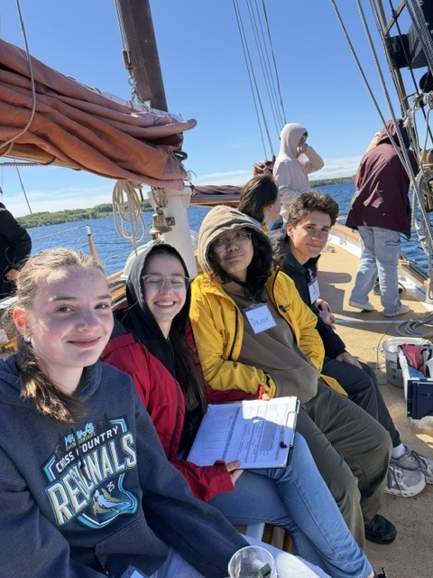 Four people sit on a boat deck, smiling, while one holds papers. A few people stand behind them.