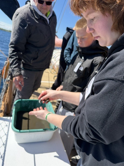 Three people on a boat, one examining an object in a container, another person watches.