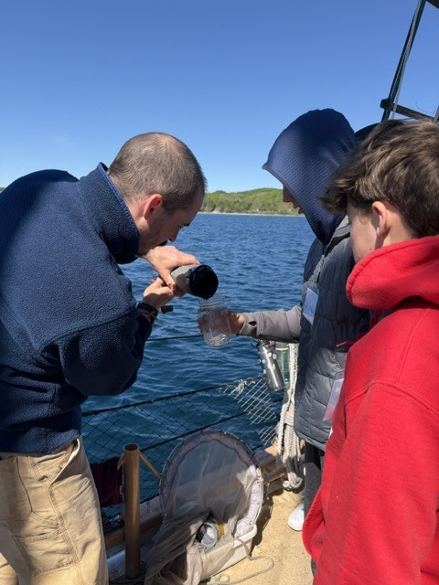 Three people examine a water sample in a clear container. One man holds a black object, possibly a device. They stand on a deck, with water and distant land visible.