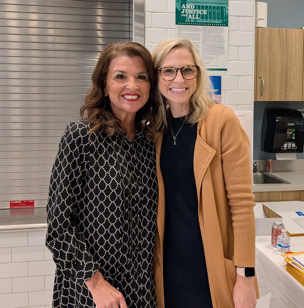 Two women stand smiling next to each other in a room with tiled walls and a white countertop.