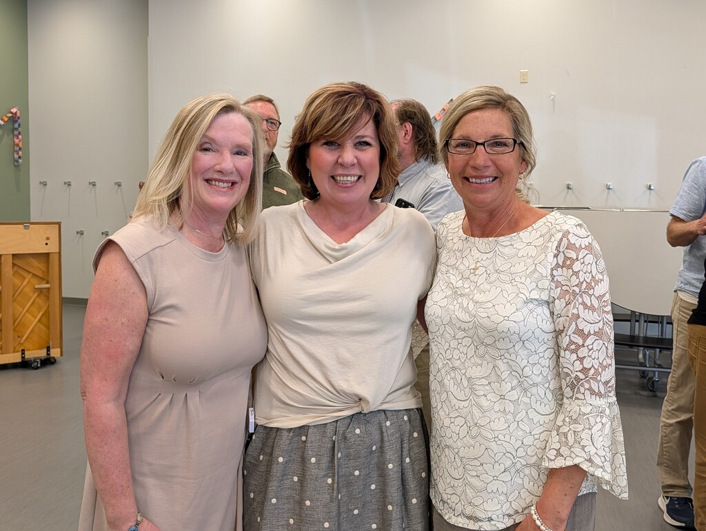 Three women stand smiling with a person in a tan shirt in the background. The background contains a wooden piano.