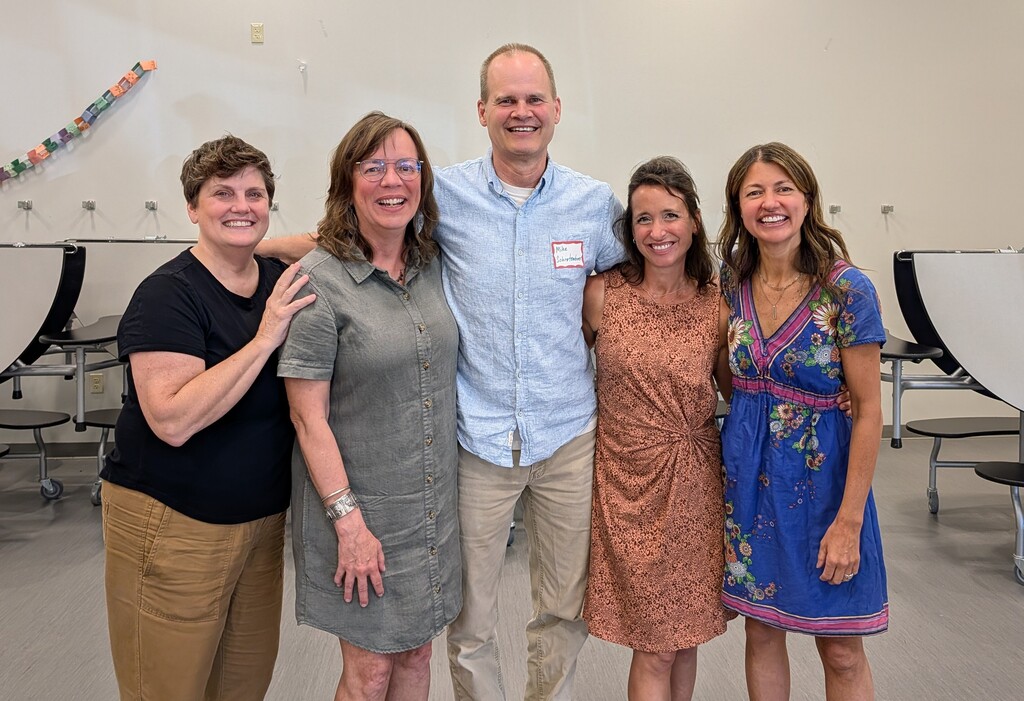 Five people in light clothing pose together in a room with white walls and chairs behind them.