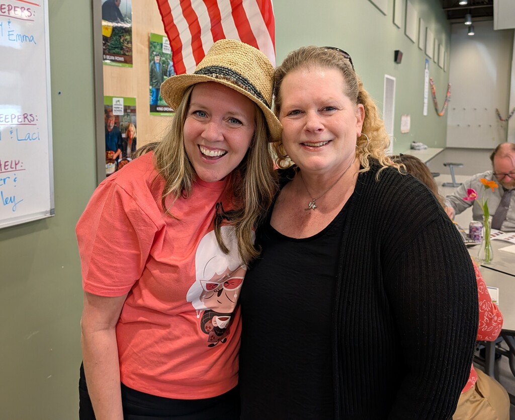 Two women pose together for a photo. One wears a straw hat and orange shirt. The other wears a black sweater.