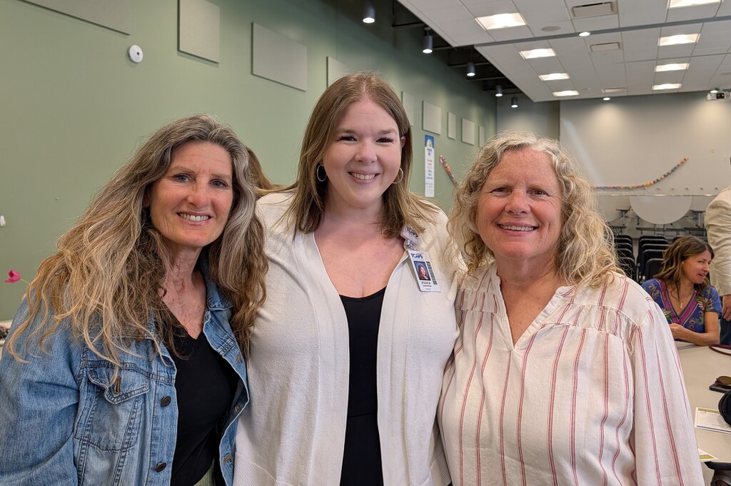 Three women stand together in a room with white walls and green accents. One wears a denim jacket, one a white blouse, and the other a striped shirt.