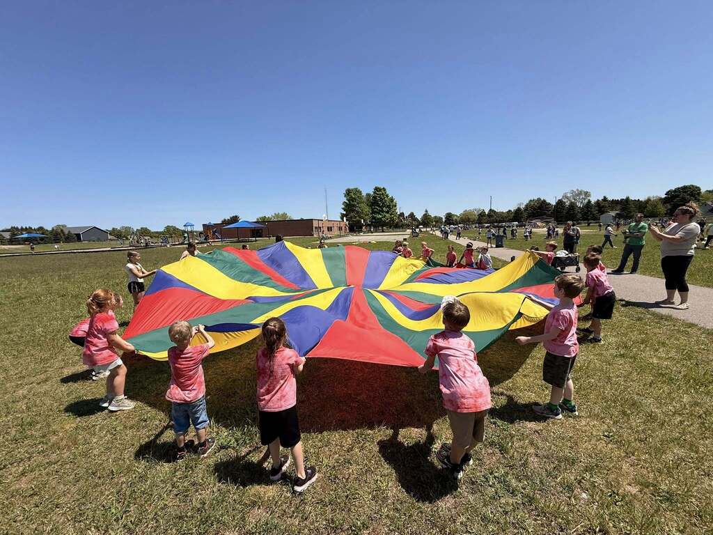 Kids in pink shirts gather around a multicolored parachute on a grassy field. People are in the background.